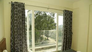 A hotel room with a large window, a bed with a white blanket, and a view of trees and a building outside. - Silicon Inn (Bengaluru)