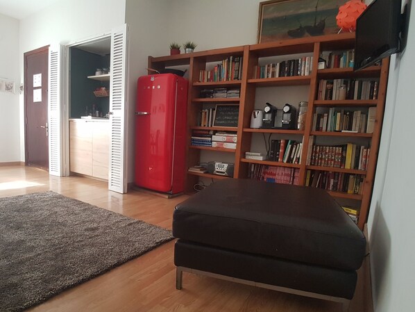A living room with a red refrigerator, a bookshelf filled with books, a grey sofa, and a wooden floor. - 2 Rooms in the center of Seville (Sevilla)