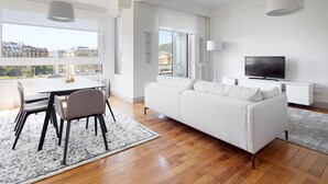 A modern living room with a white sofa, dining table, and chairs. There is a TV on a white cabinet, and large windows offer a city view. - Okendo by FeelFree Rentals (San Sebastián)