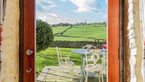 A view from a room with a wooden door leading to a patio with a table and chairs, a stone wall, and a green field with a power line in the distance. - The Manger, Perched in an elevated position within the Blackdown Hills National Landscape. (Axminster)