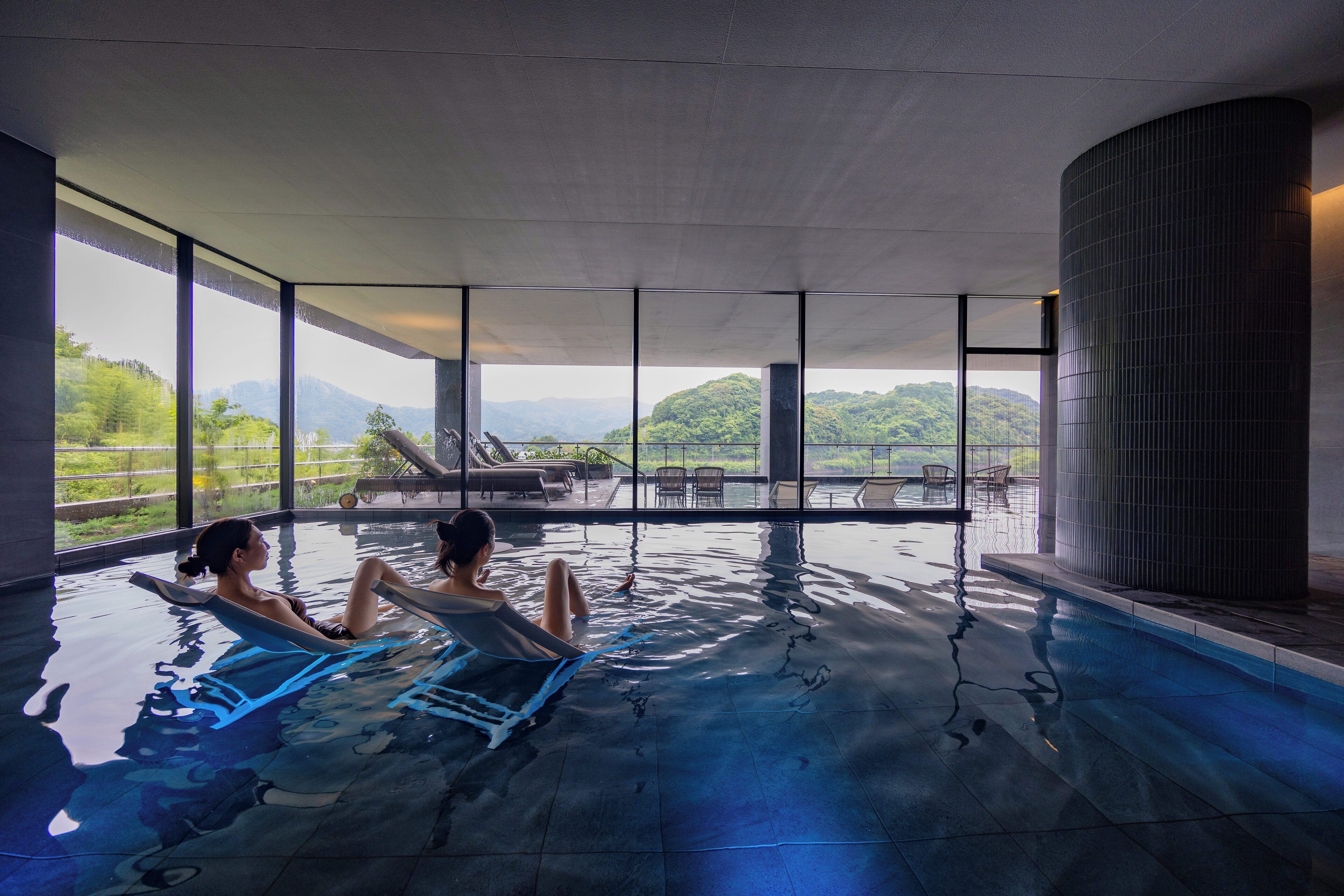 Una piscina interior con dos personas relajándose en sillones de descanso, ventanas amplias y vistas a montañas.