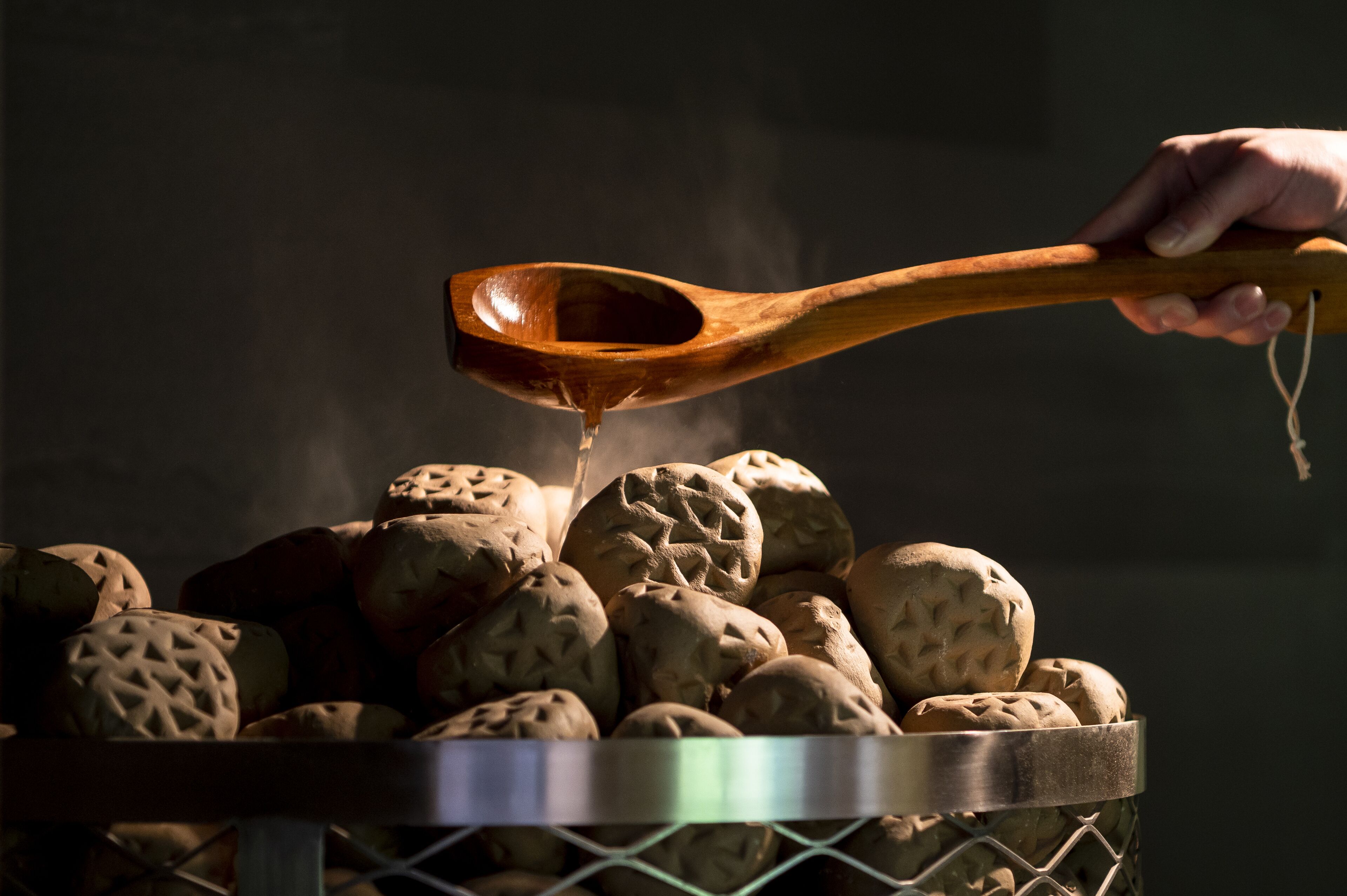 A hand holding a wooden spoon over a basket filled with round, textured items, likely food, with steam rising from the basket.