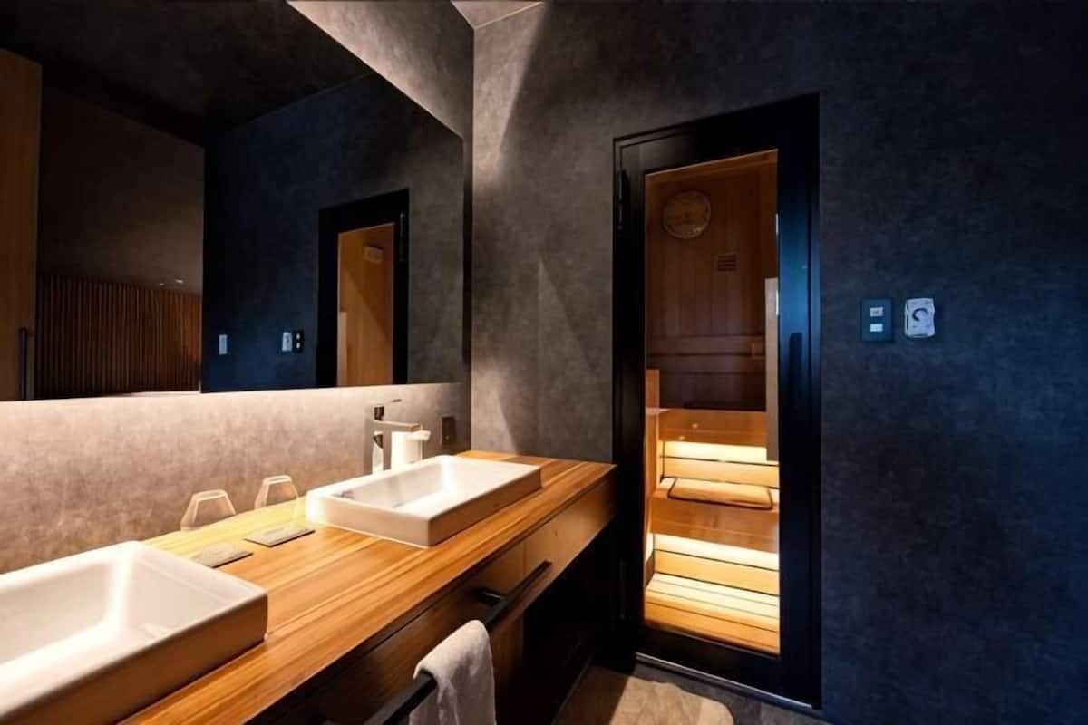 A modern bathroom with two sinks, a wooden vanity, a large mirror, and a sauna visible through a door.