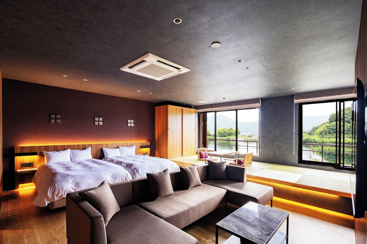 A modern hotel room with a large bed, a sofa, a small table, and a view of a lake.