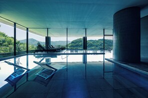 Ein moderner, überdachter Pool mit großen Glaswänden, Liegestühlen und Blick auf eine Berglandschaft.