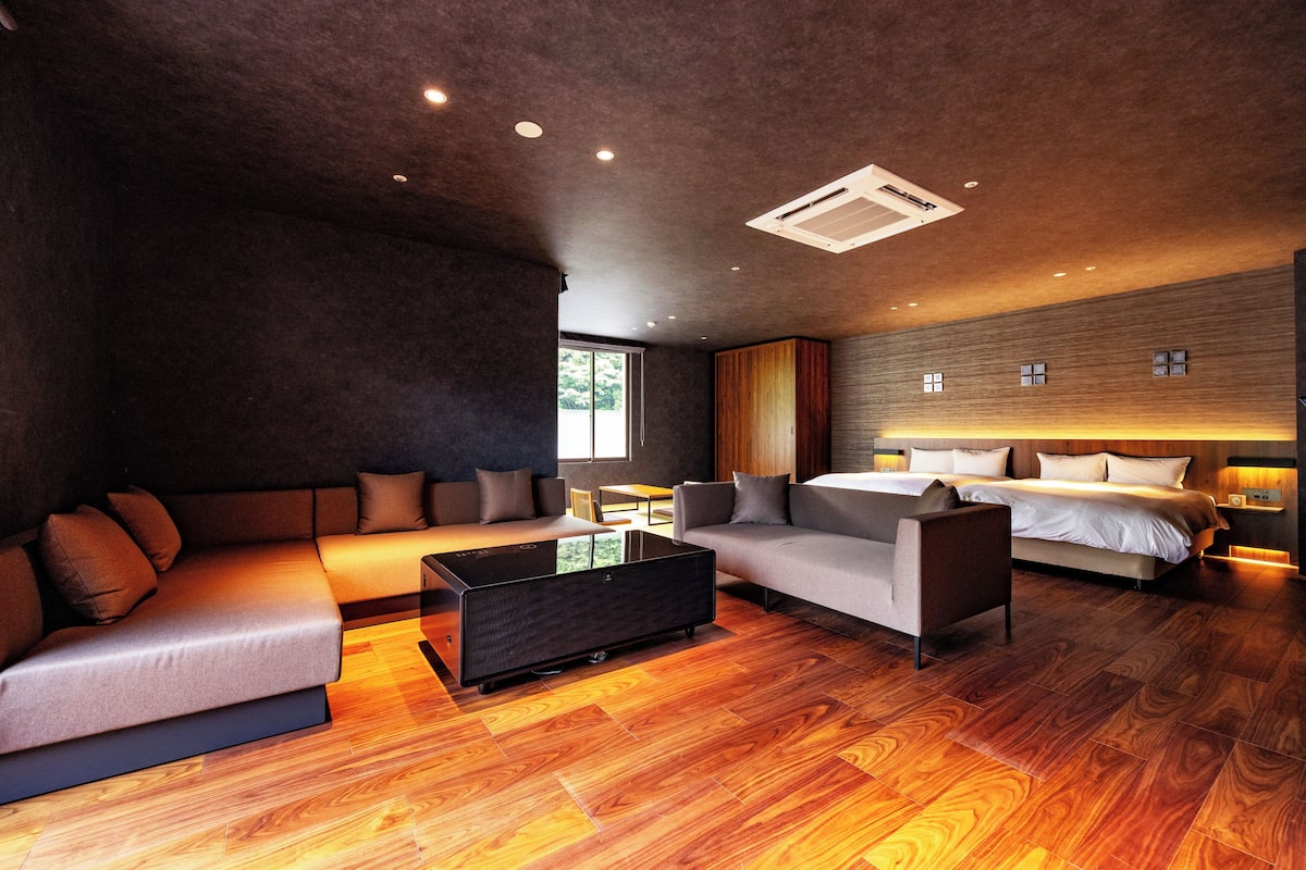 A modern hotel room with a sectional sofa, a small table, a bed, and a wooden floor.