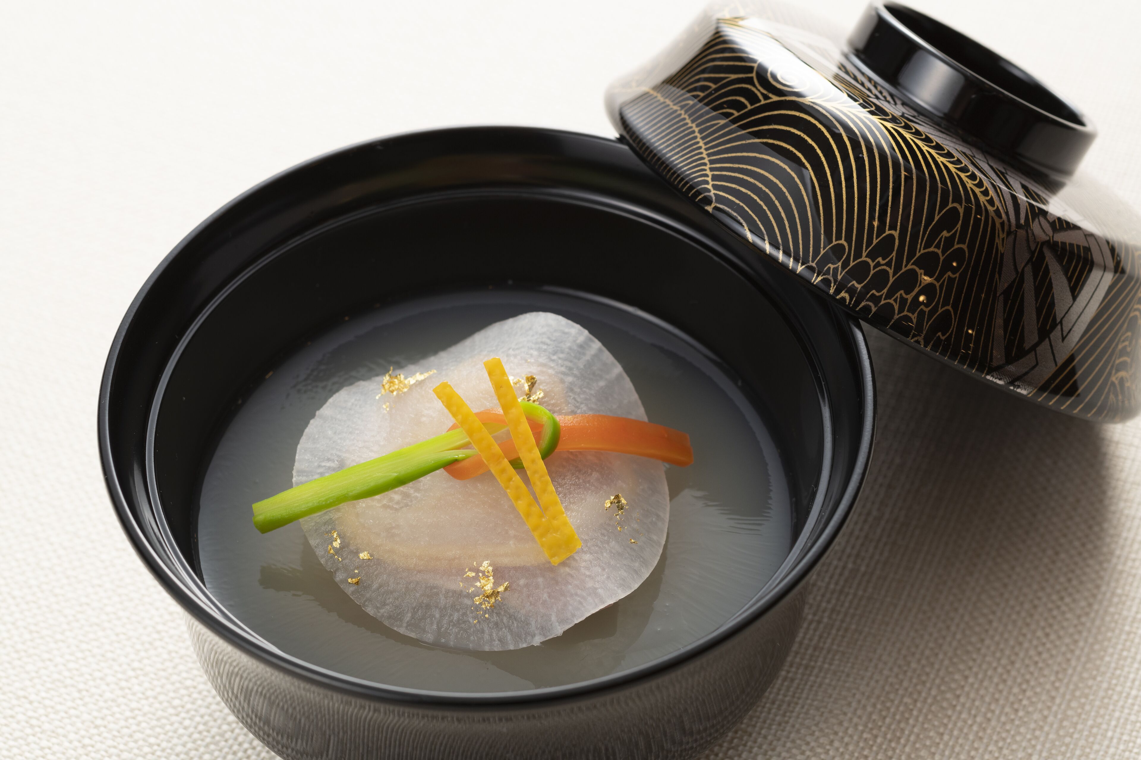 Un tazón de sopa transparente con una porción de pescado y adornos, acompañado de un recipiente decorativo.
