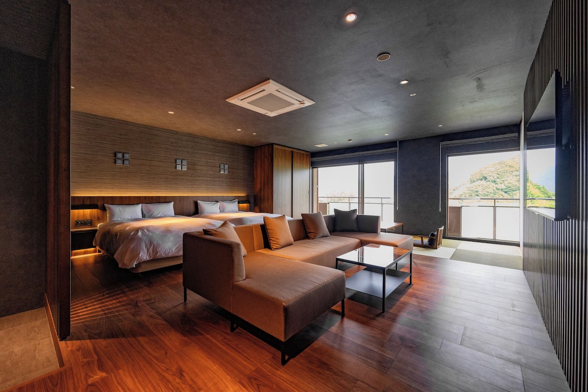 A modern hotel room with a large bed, a sofa, a coffee table, and a view of the outdoors.