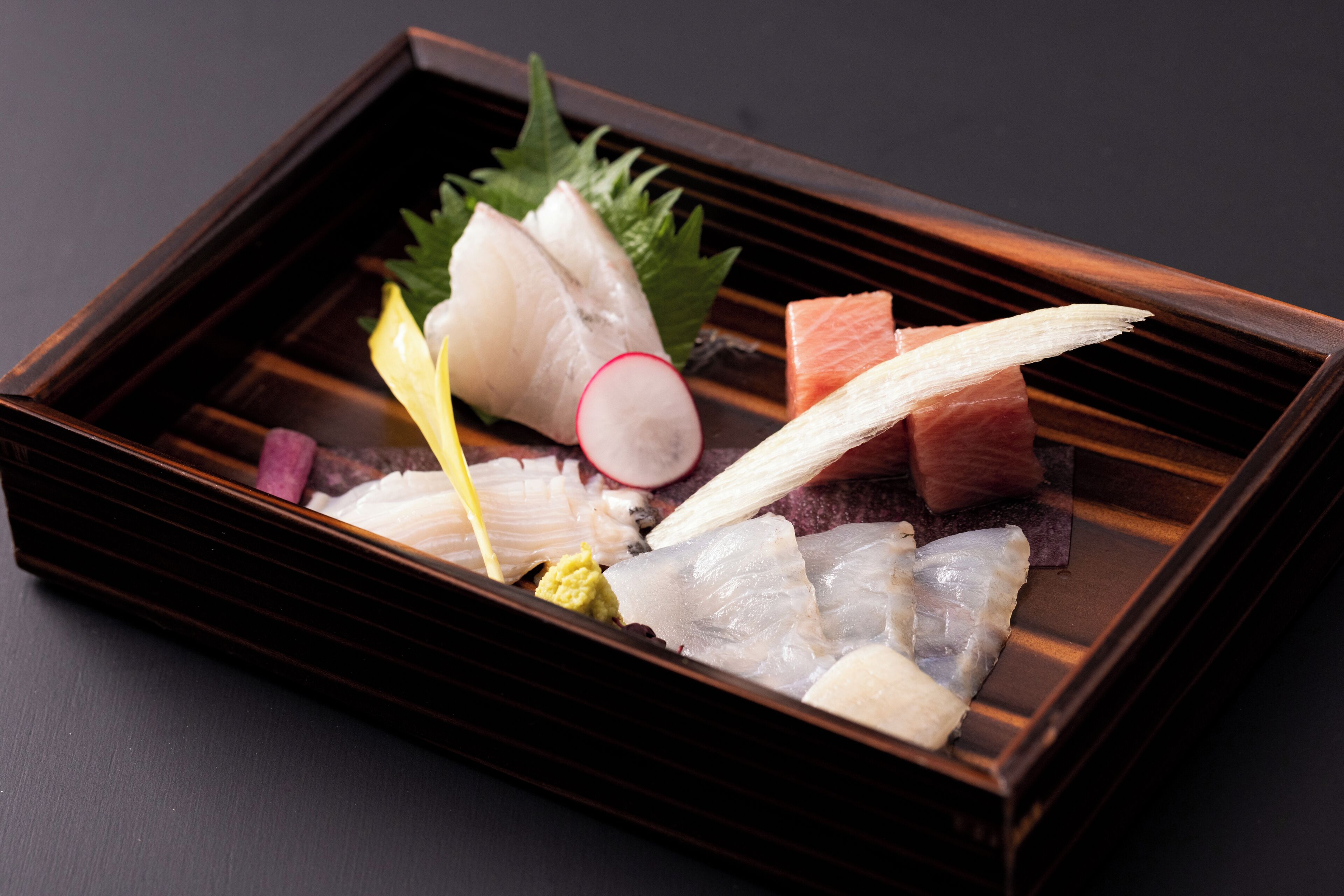 Ein Holztablett mit verschiedenen Meeresfrüchten, darunter Fisch und möglicherweise Fischrogen, auf einer dunklen Oberfläche.