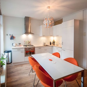 Moderni keittiö, jossa on valkoiset kaapit, ruostumattomasta teräksestä valmistettu liesituuletin, kaasuliesi ja ruokapöytä, jonka ympärillä on oranssit tuolit.