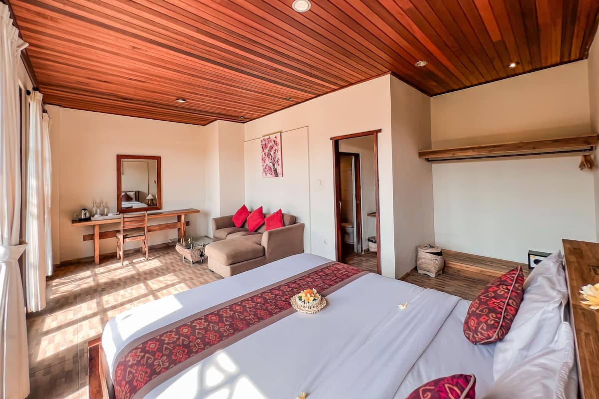 Un dormitorio con techo de madera, una cama con un corredor estampado, un escritorio, un sofá con cojines rojos y un baño visible a través de una puerta abierta.