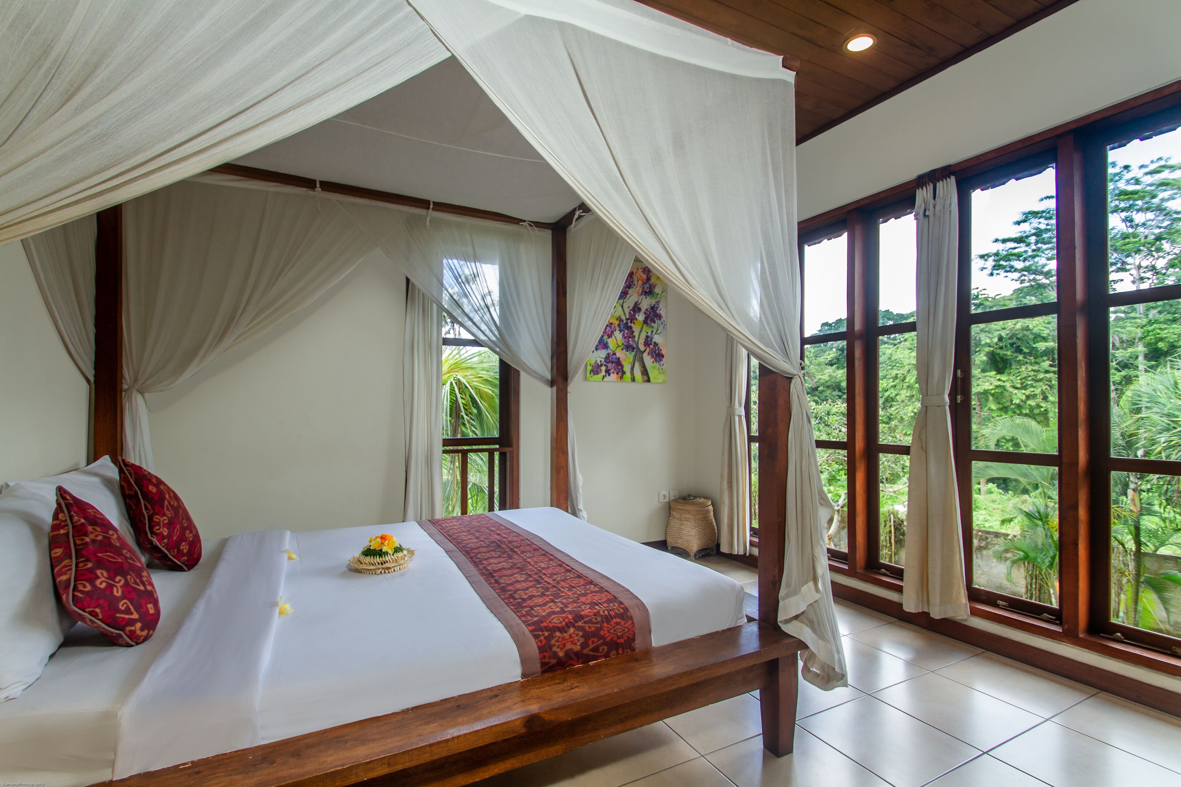 A four-poster bed with a canopy, a large window with curtains, and a tropical view.