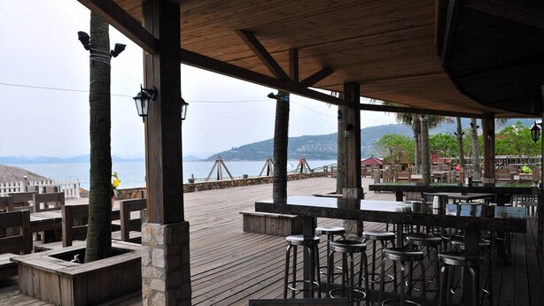 A wooden deck with stone pillars, bar stools, and tables overlooking a body of water with a distant mountain view. - Da Mei Sha Yacht Club Resort (Shenzhen)