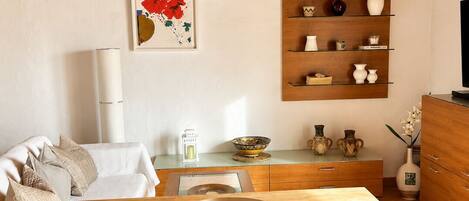 A dining area with a wooden table, white chairs, a white sofa, a wall-mounted shelf with decorative items, and a framed artwork.