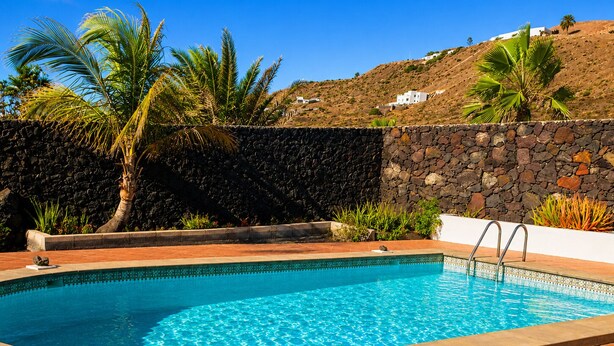 A rectangular swimming pool with clear blue water, surrounded by a stone wall and palm trees. - Wonderful views of the sea and the volcanoes. Casa Isabel  (Tías)