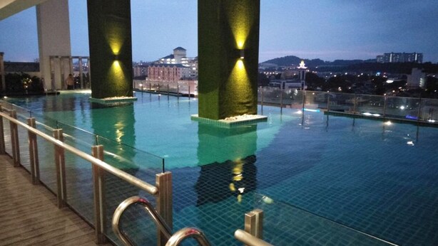 An indoor infinity pool with clear glass railings, large pillars, and city views at dusk. - Elegant Cube Soho One South (Seri Kembangan)