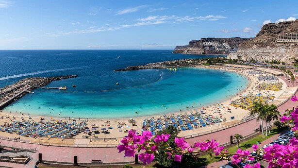 A coastal beach with a curved shoreline, a promenade lined with umbrellas, and a rocky cliff in the background.