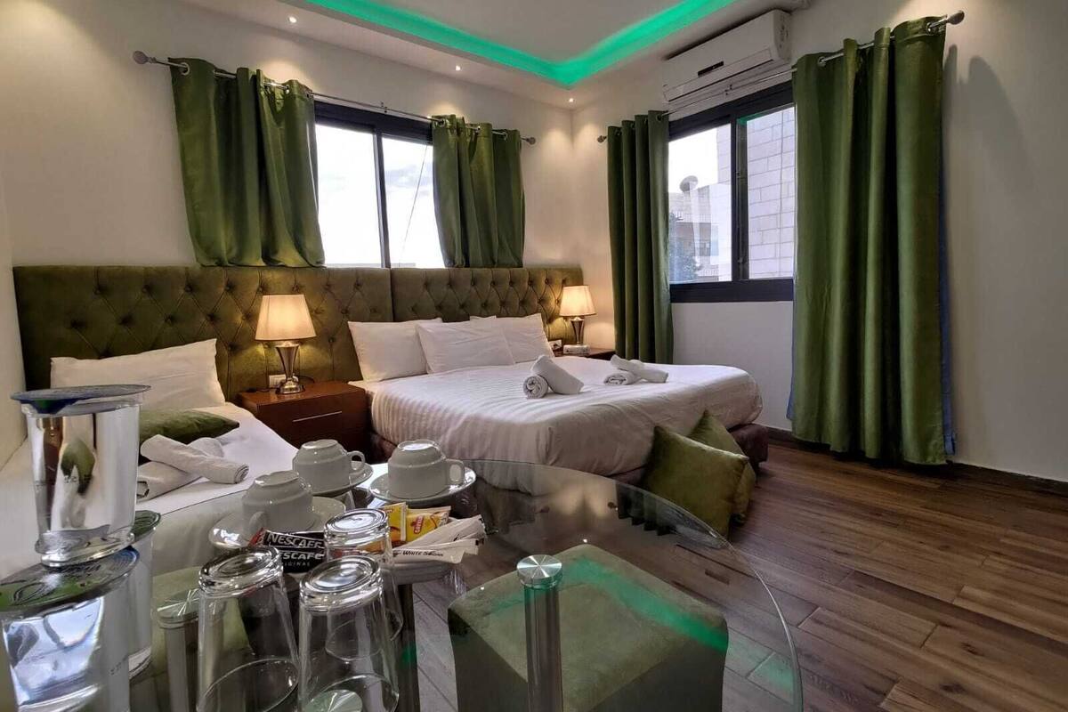 A hotel room with a large bed, two bedside tables, a desk, and a chair. There are curtains, a window, and an air conditioning unit.