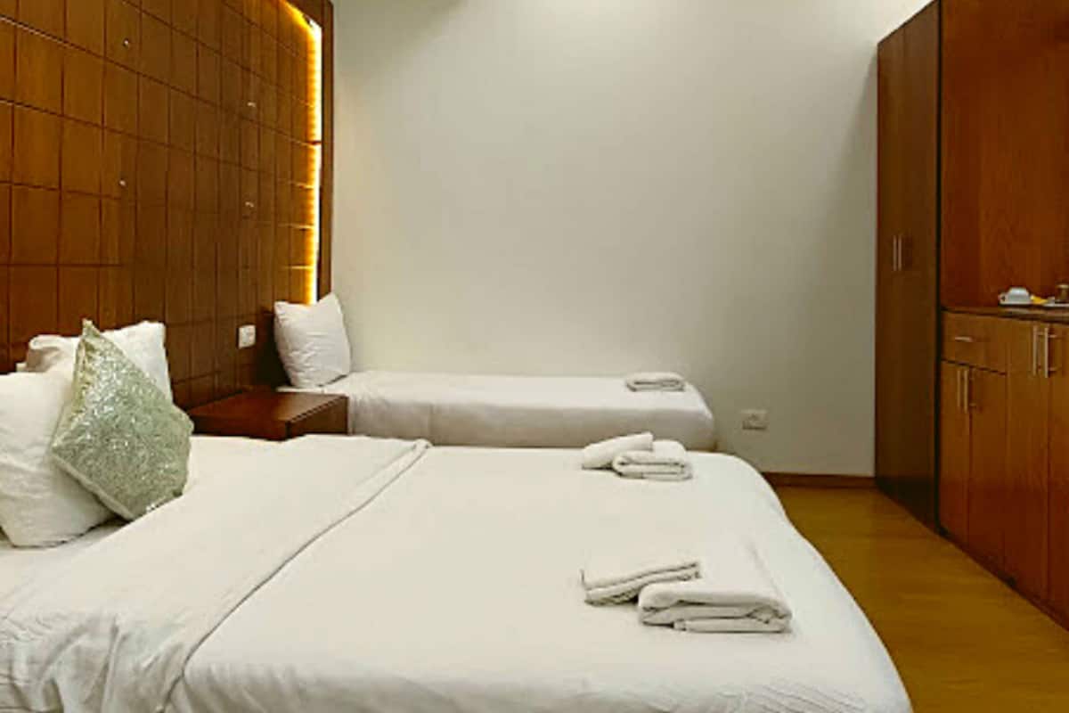 A hotel room with two beds, a wooden wardrobe, a grey armchair, and a small table with a kettle.
