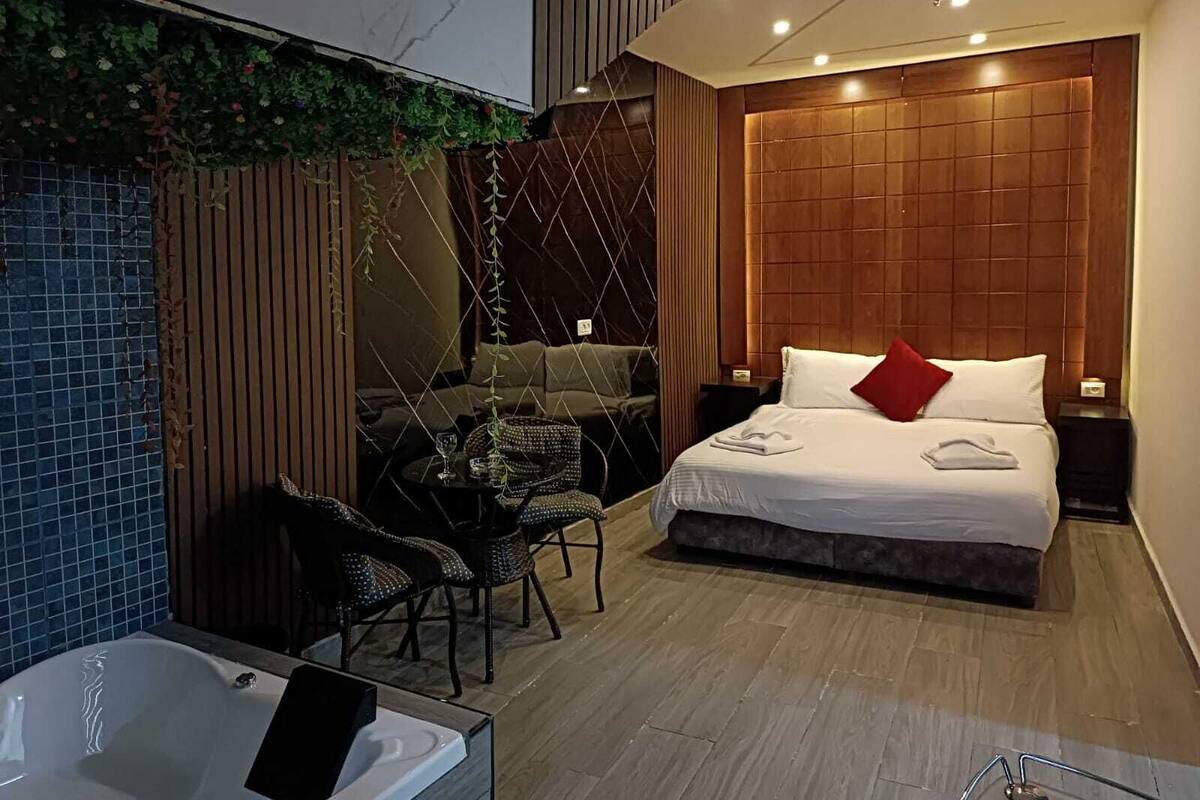 A modern hotel room with a large bed, a sitting area with a sofa and chairs, a small table, and a bathtub.