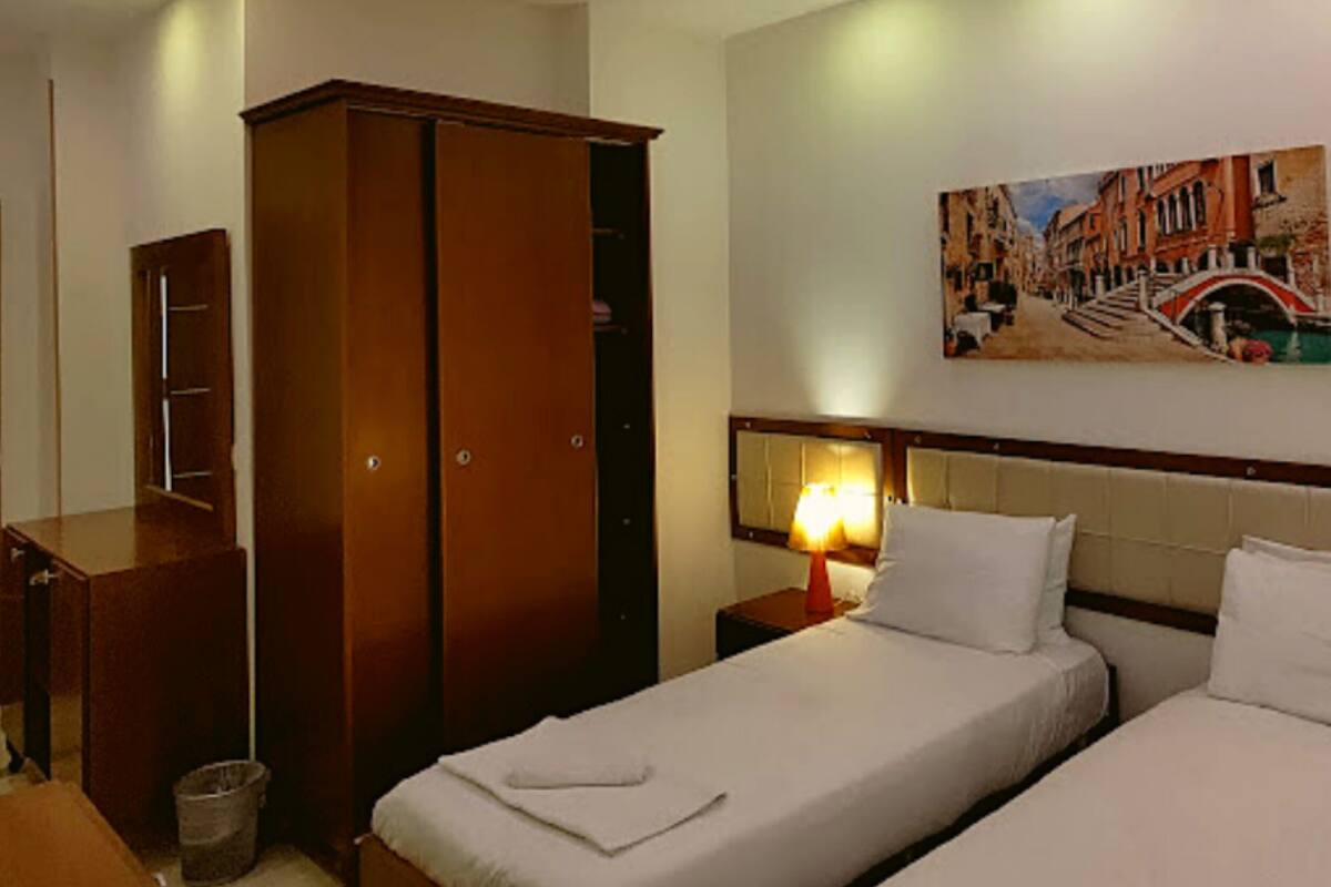 A hotel room with two single beds, a wardrobe, a painting of a cityscape, and a nightstand with a lamp.