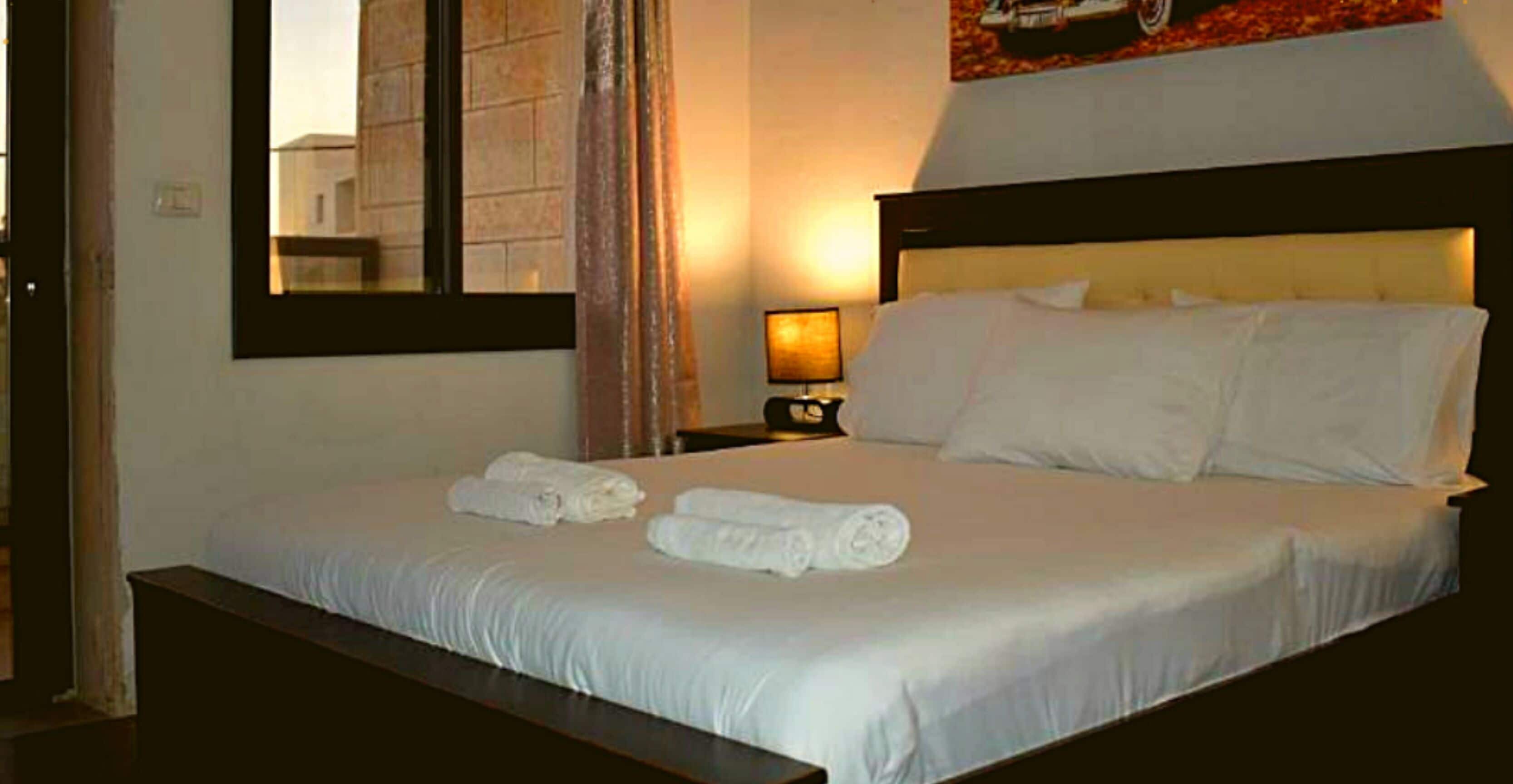 A neatly made bed with white linens and towels, a bedside lamp, and a framed picture on the wall.