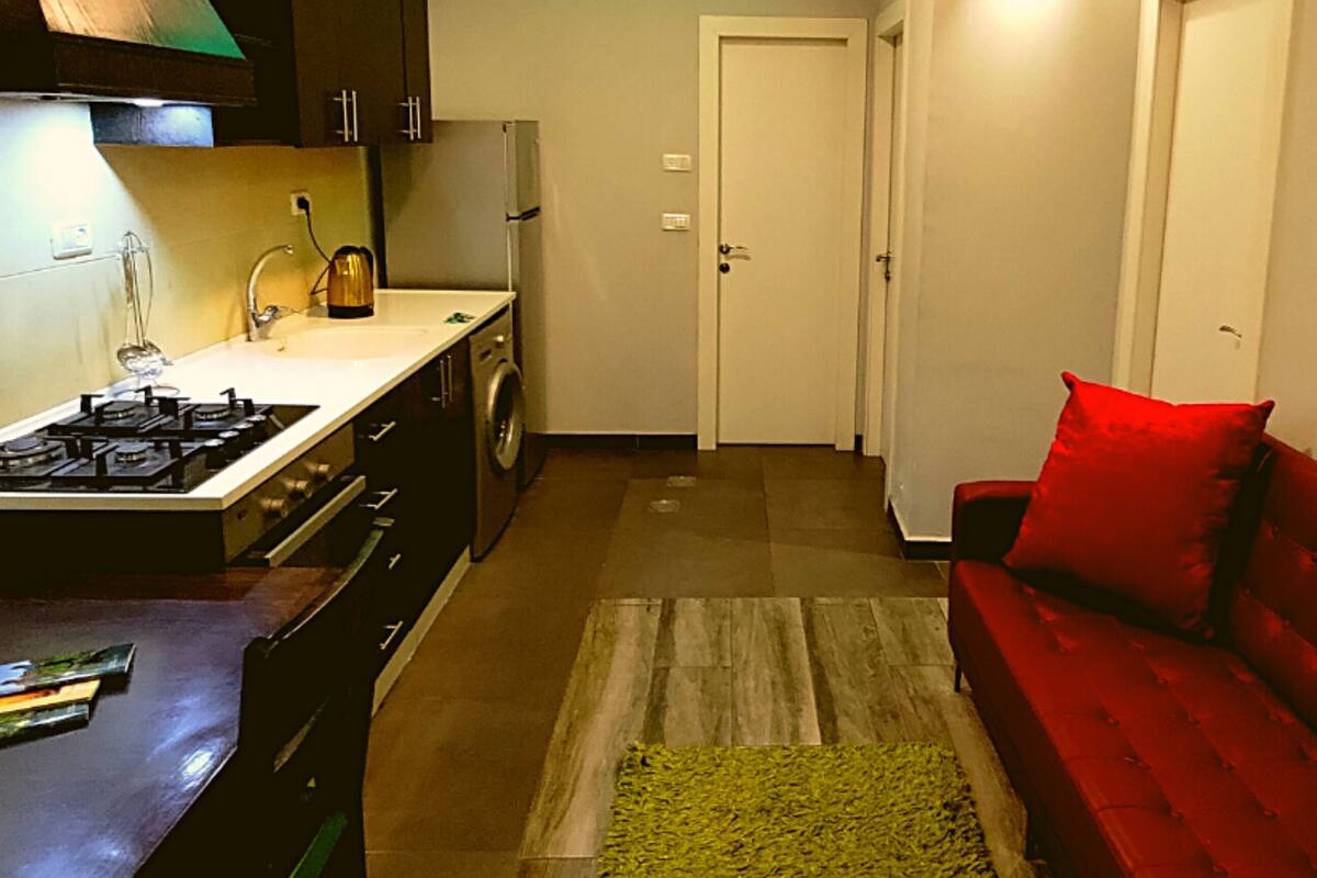 A compact kitchen with a gas stove, a washing machine, and a red sofa in a narrow hallway.