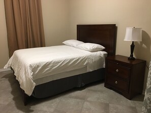 Un letto rifatto con lenzuola bianche, un comodino in legno con una lampada e una tenda marrone.