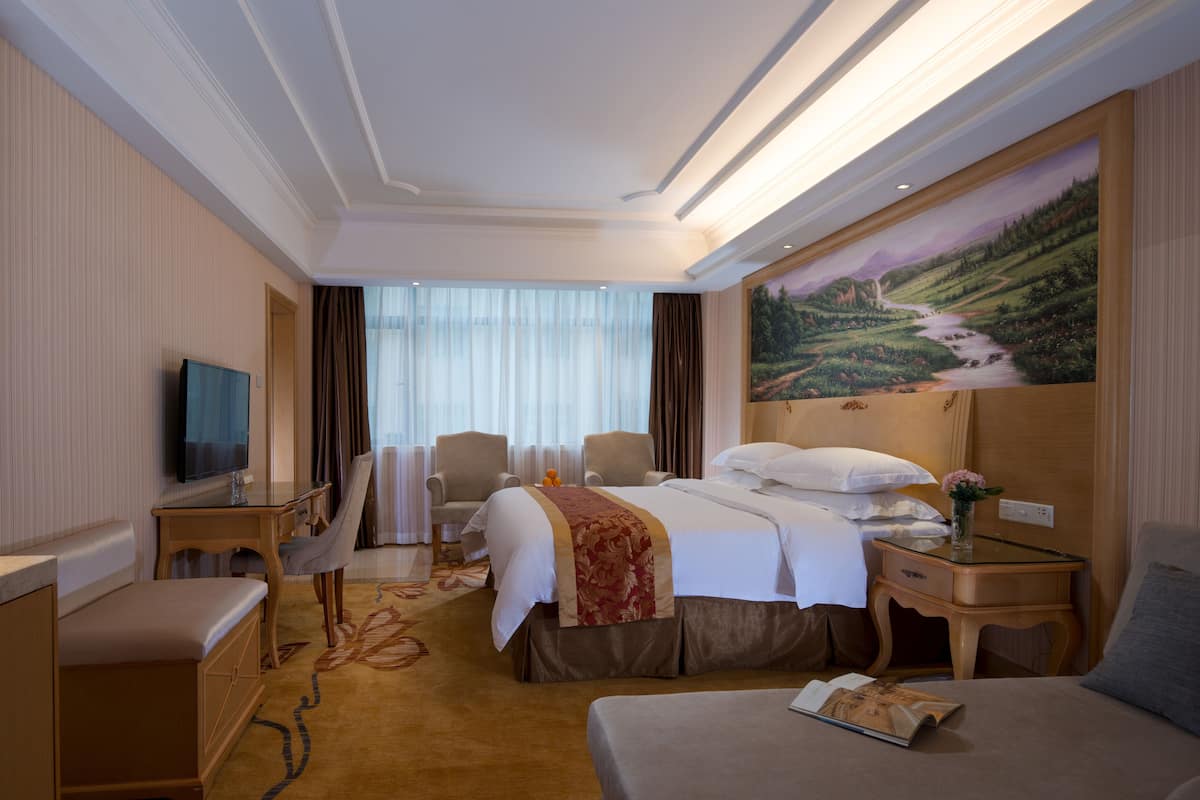 A hotel room with a bed, a desk, a TV, a chair, a sofa, and a painting on the wall.