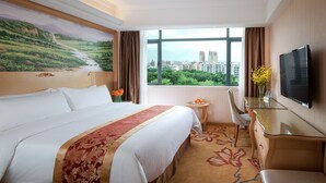 A hotel room with a large bed, a desk with a TV, a small table with fruit, and a window with a city view. - Vienna Hotel -Yantian Harbor (Shenzhen)