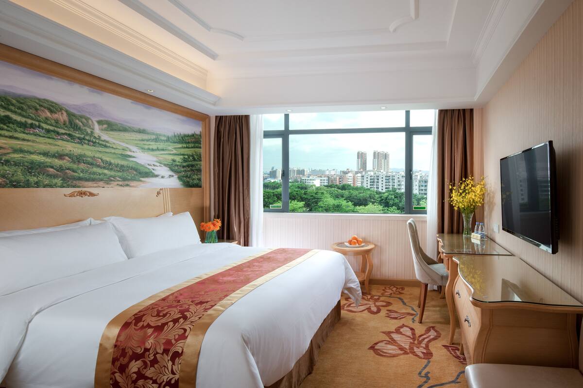 A hotel room with a large bed, a desk with a TV, a small table with fruit, and a window with a city view.