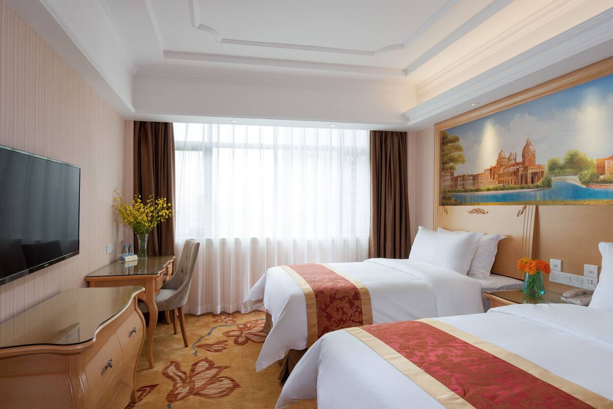 A hotel room with two beds, a TV, a desk, and a painting of a historic building.
