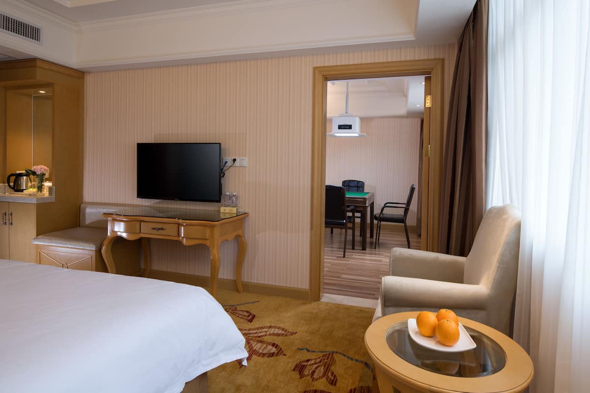 A hotel room with a bed, a desk with a TV, a chair, a small table with fruit, and a view of another room through an open door.