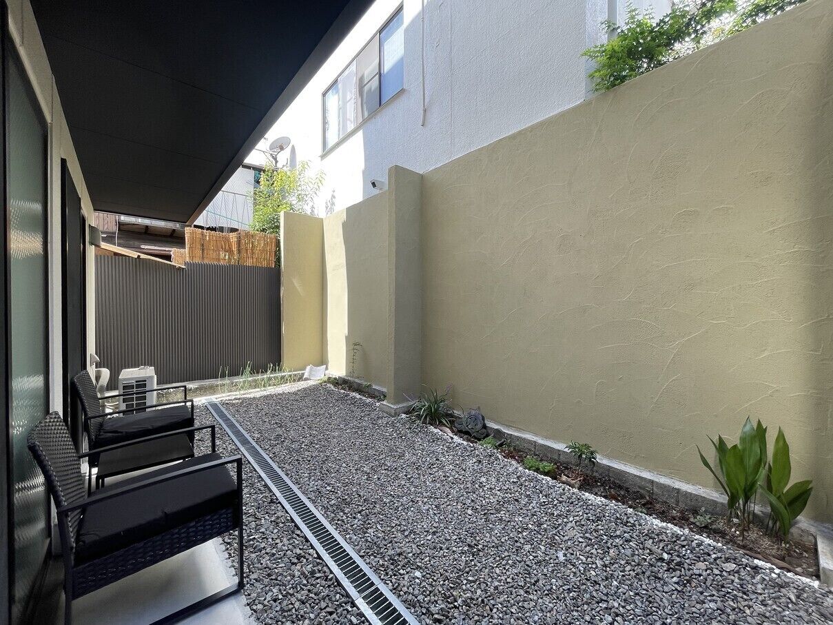 A modern outdoor seating area with a metal grill, a small garden with plants, and a building with a beige wall and a window.