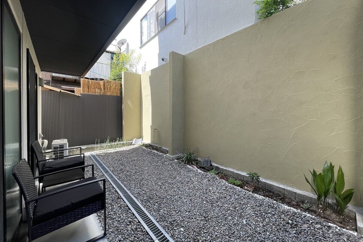 A modern outdoor seating area with a metal grill, a small garden with plants, and a building with a beige wall and a window.