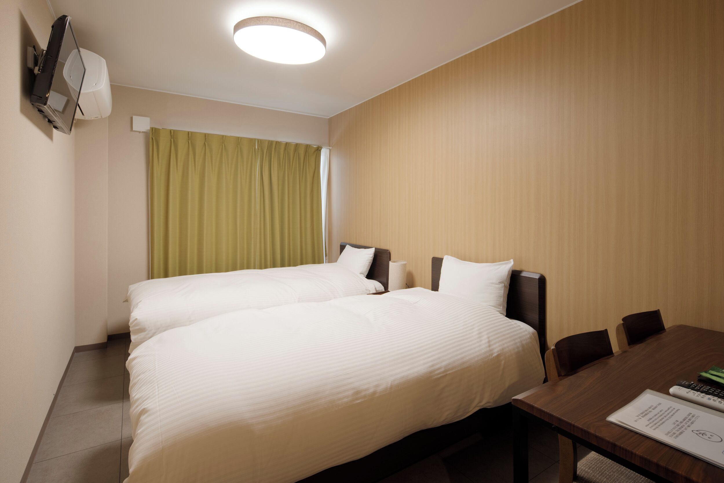 A hotel room with two beds, a wooden wall, a table with chairs, and a ceiling light.