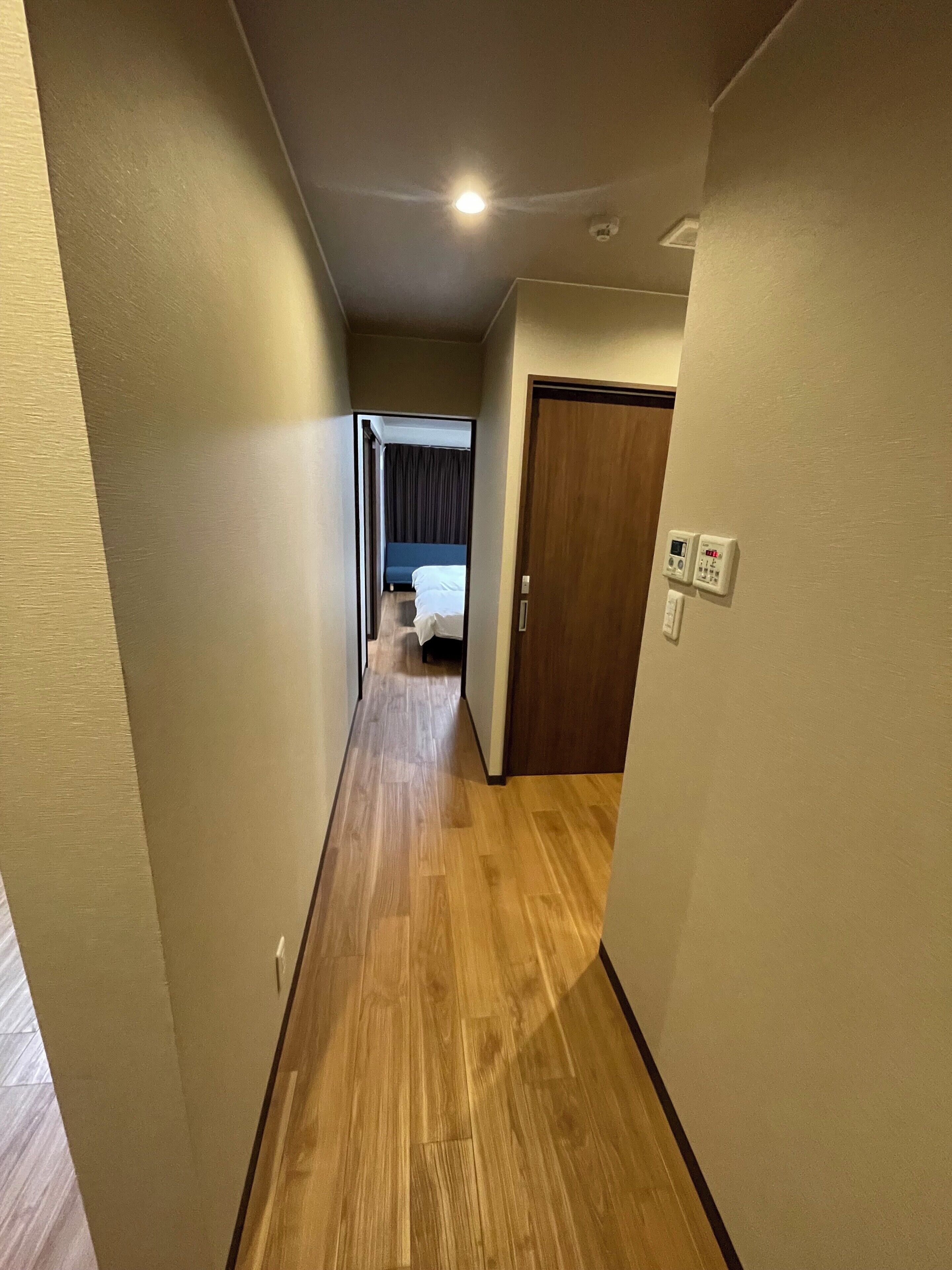 Un couloir étroit, doté d’un plancher en bois, mène à une pièce où se trouvent un lit et une porte.
