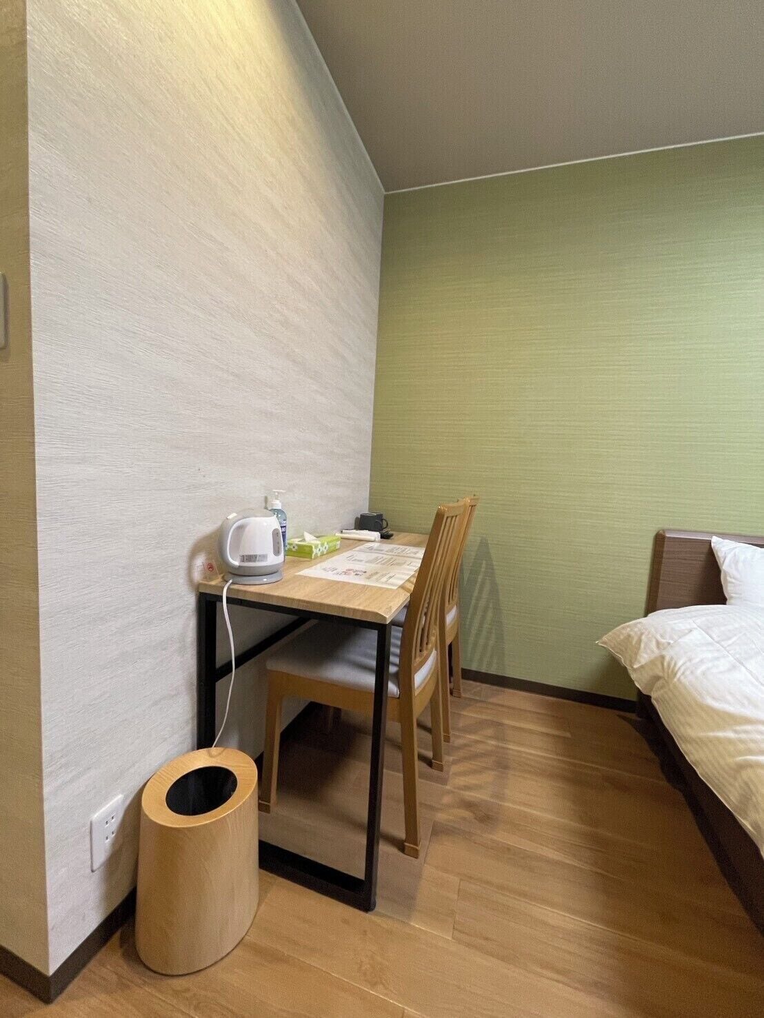 A small, neatly arranged room with a bed, a table with a chair, a waste bin, and a wall-mounted heater.