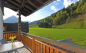 Un balcón con barandilla de madera, una mesa con un florero y vistas a un campo verde y colinas boscosas.