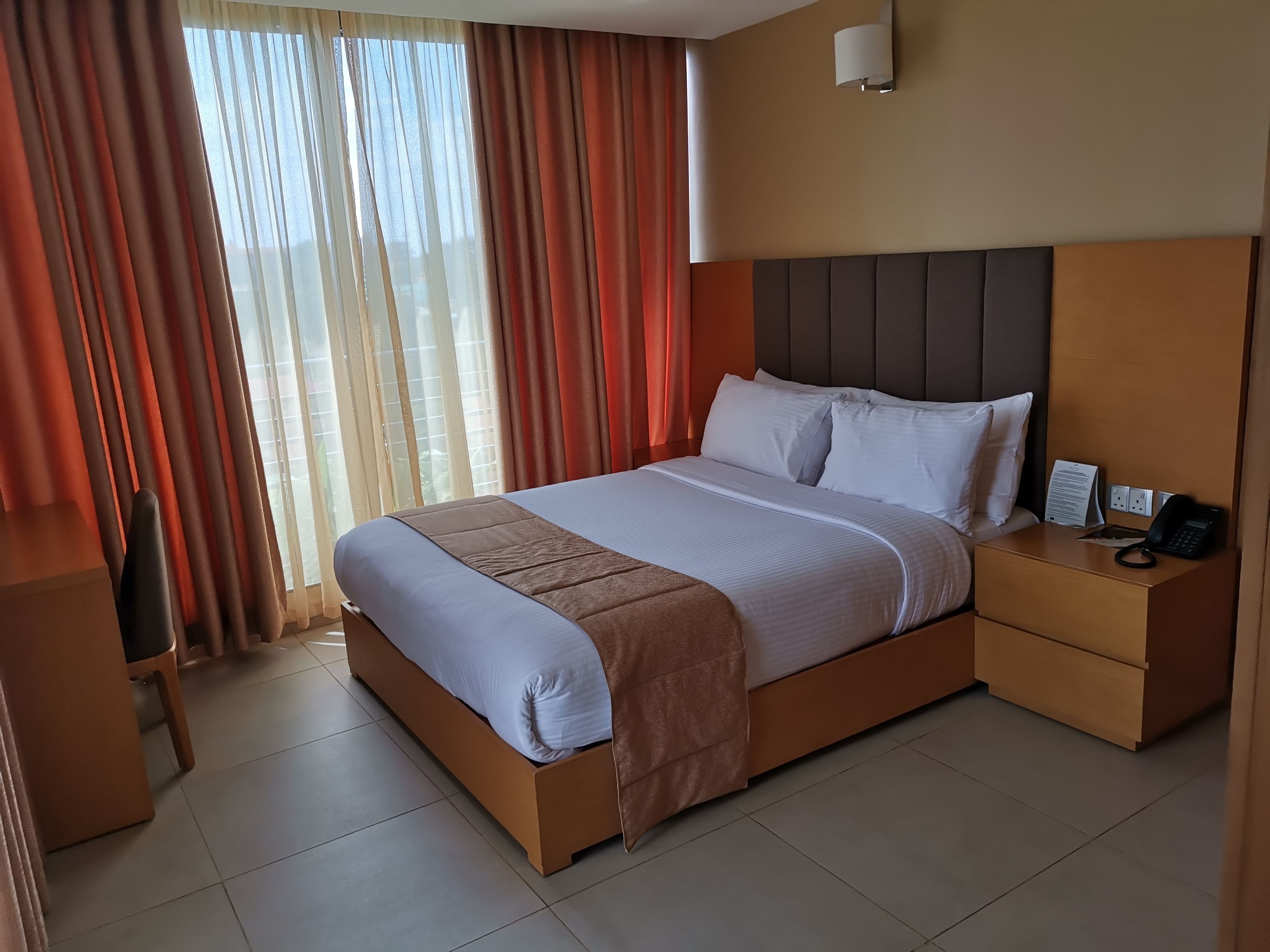 Een hotelkamer met een groot bed, een nachtkastje met een telefoon, en een uitzicht door dunne gordijnen.
