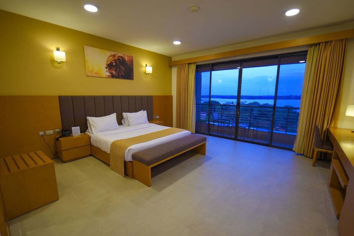 Habitación de hotel con una cama grande, una mesita de noche, un banco, un escritorio y vistas de la ciudad a través de un ventanal amplio.
