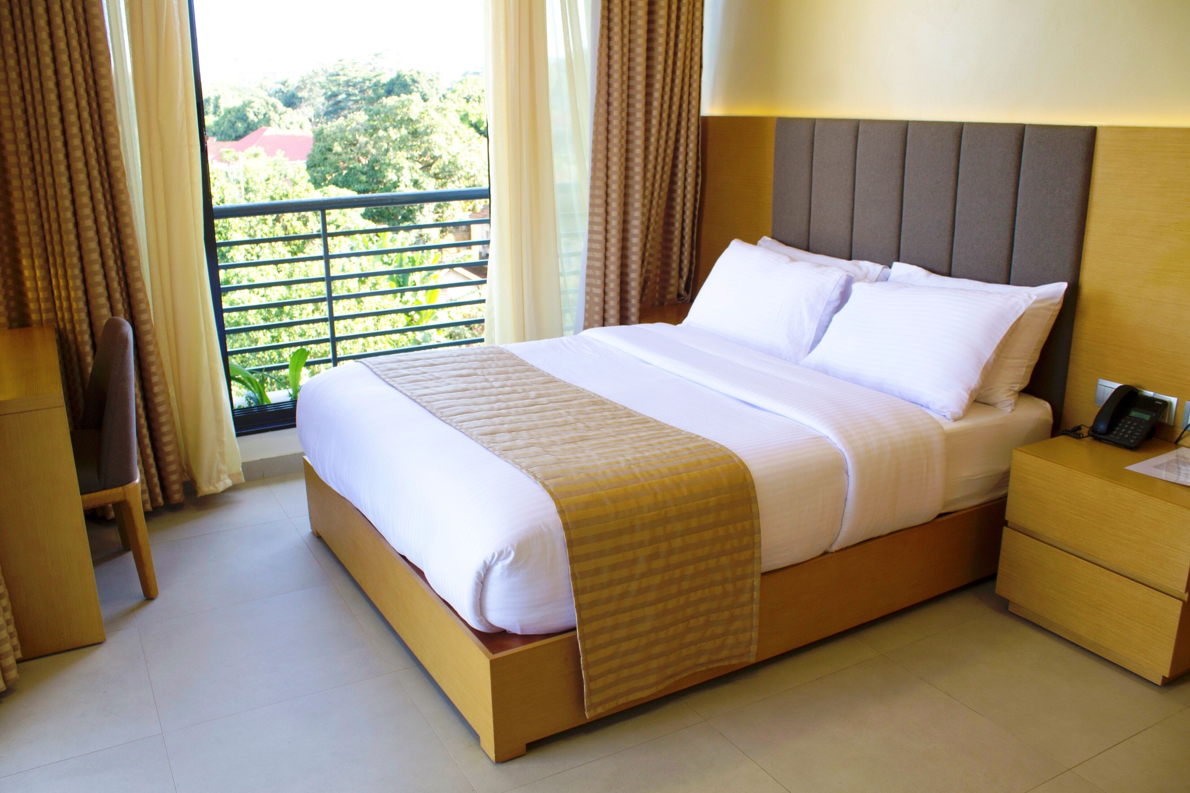 A hotel room with a large bed, a bedside table with a phone, and a view of greenery outside.