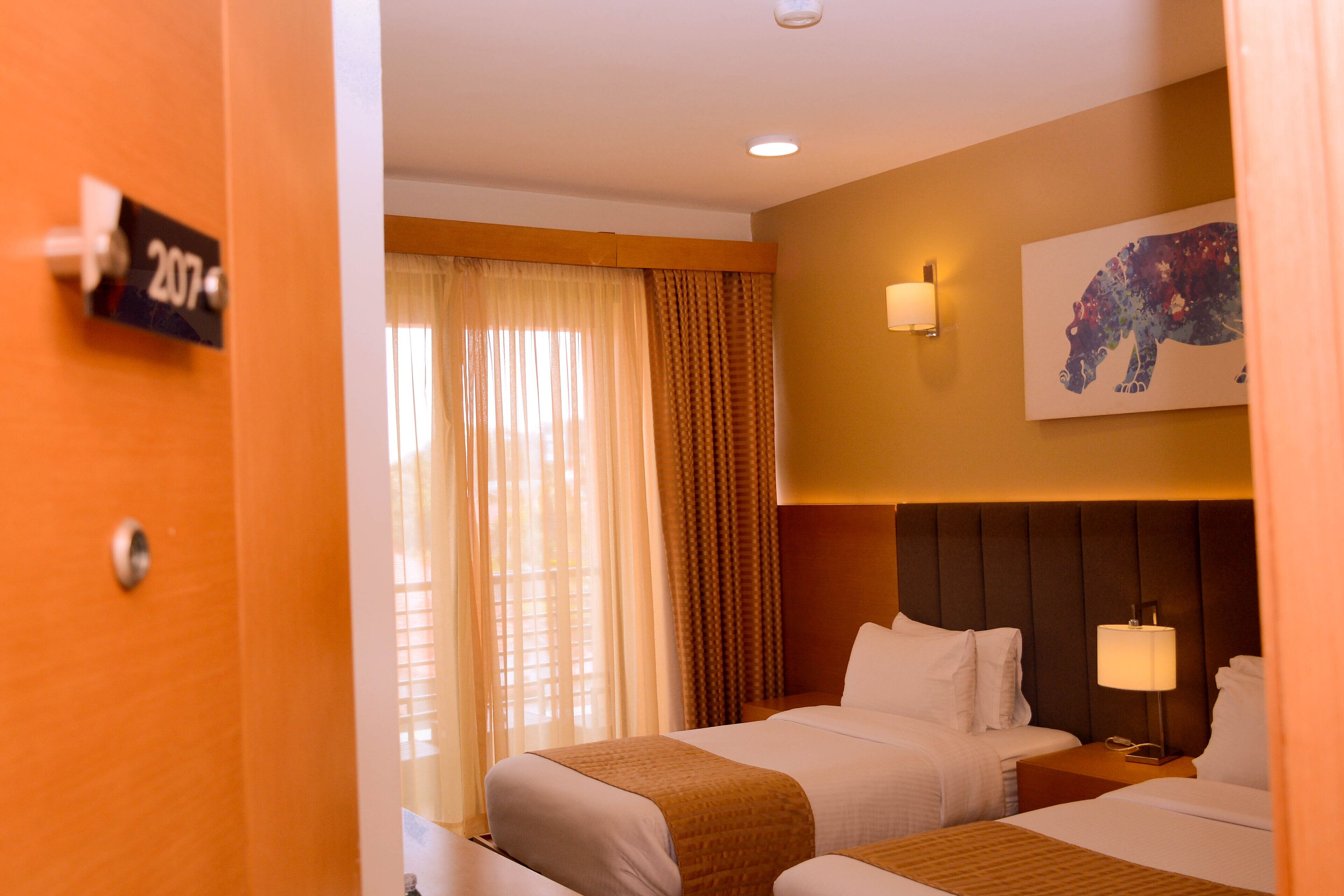 A hotel room with two beds, a large window with curtains, a wall-mounted lamp, and a framed animal picture.