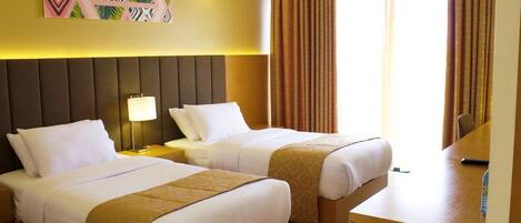 A hotel room with two beds, a desk, a lamp, and a painting on the wall.