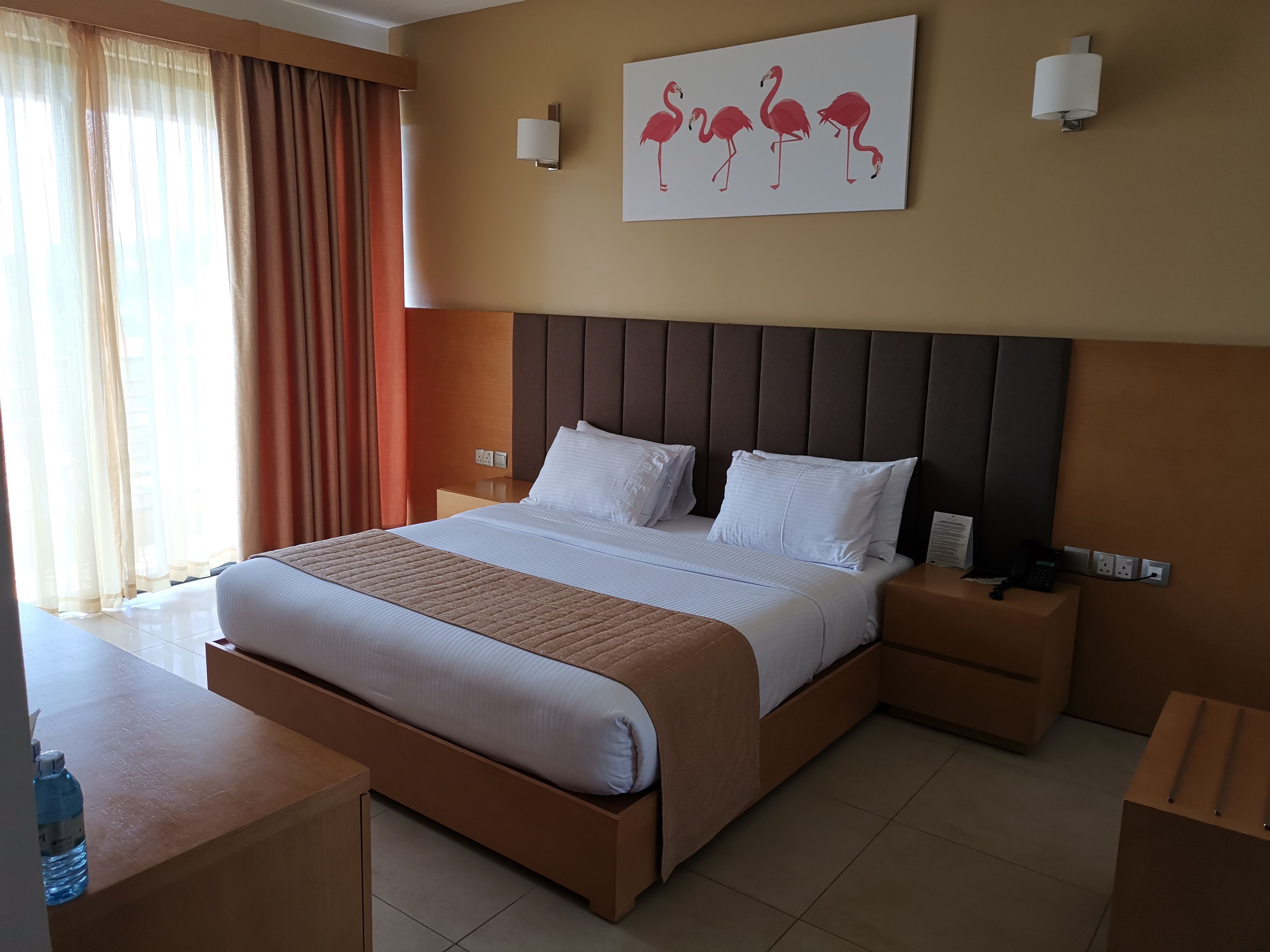 A hotel room with a large bed, bedside tables, a water bottle, and a painting of flamingos on the wall.