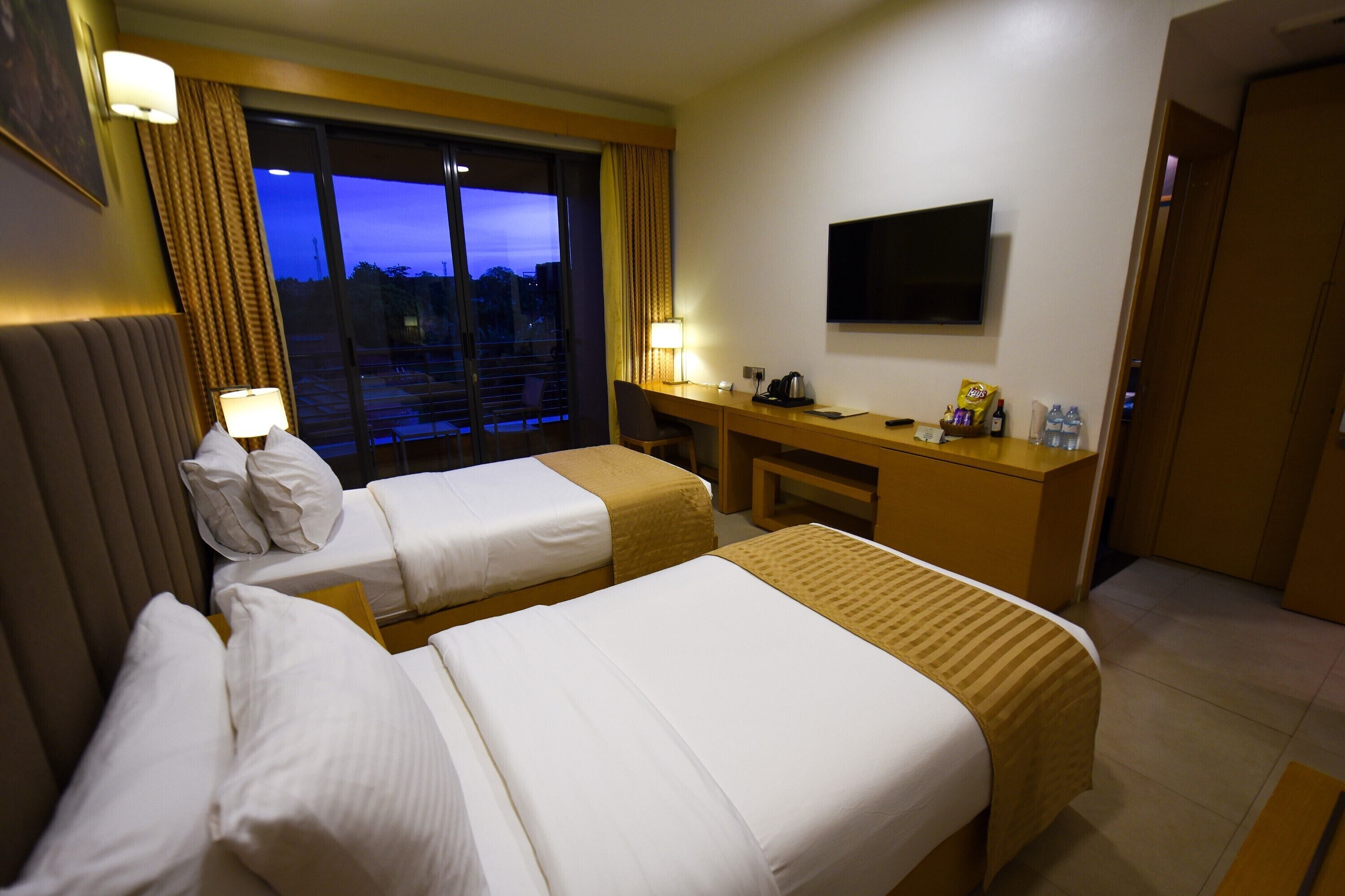 A hotel room with two beds, a desk, a TV, and a window with a view of the city at dusk.