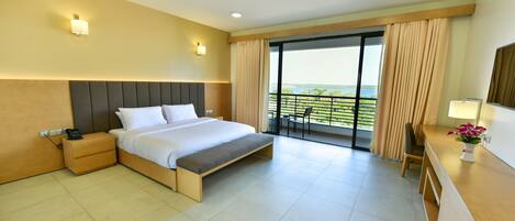 A modern hotel room with a large bed, bedside table, and a desk with a chair. The room has a balcony with a view of the outdoors.