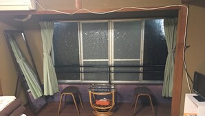 Ein Zimmer mit einem großen Fenster, zwei Holzschemeln, einem kleinen Tisch und einem Boden aus Tatami-Matten.