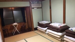 Ein Zimmer mit Tatami-Matten, einem kleinen Tisch und zwei Stühlen. Es gibt drei Betten mit Kissen und Decken. Eine Klimaanlage ist an der Wand angebracht.