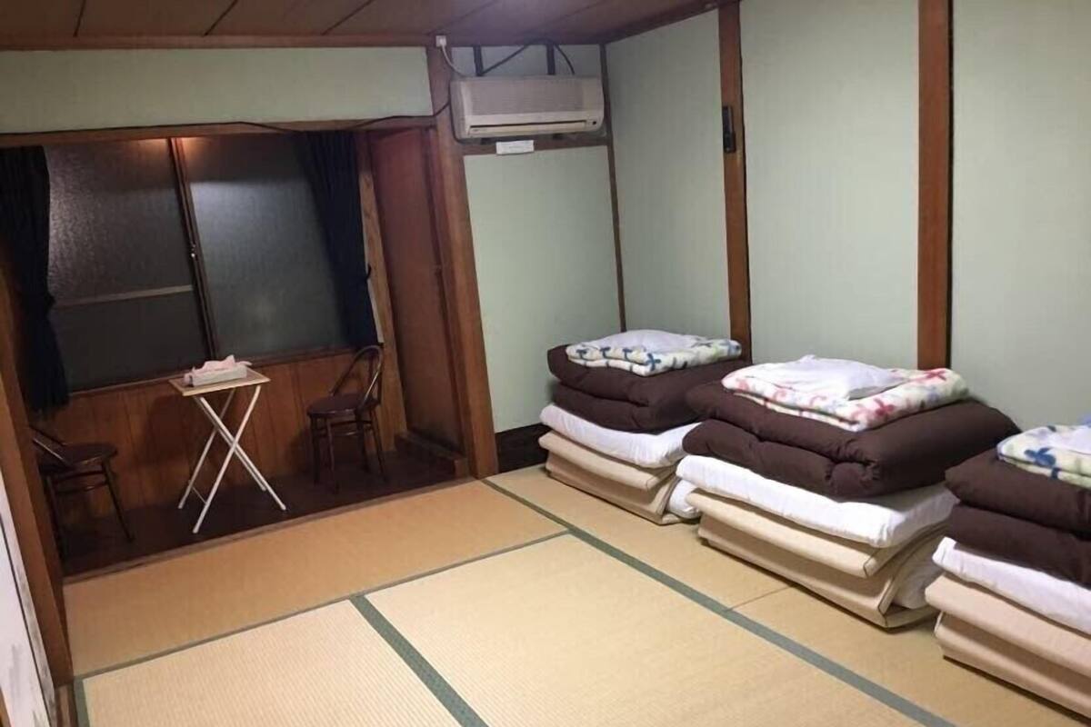 A room with tatami mats, a small table, and two chairs. There are three beds with pillows and blankets. An air conditioning unit is mounted on the wall.